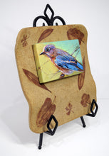Load image into Gallery viewer, Backyard Bluebird painting by Pat Cross mounted in a handbuilt and kiln fired stoneware frame resting on a black metal easel facing right.