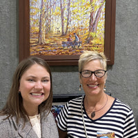 Tamarack Gallery Manager Gretchen Carr with artist Pat Cross in front of the original oil painting titled Autumn Cinema by Pat Cross.
